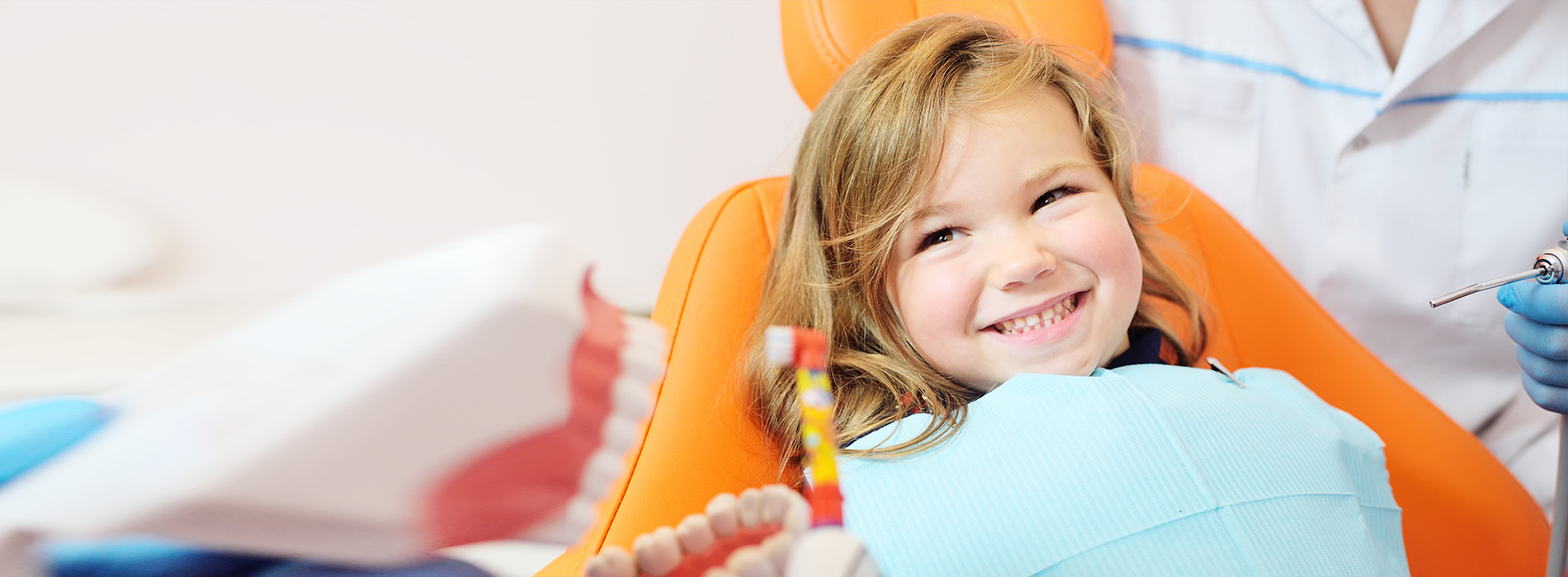 A young girl in an orange chair is smiling at the camera, with a dental professional seated behind her, ready to perform an examination or procedure.