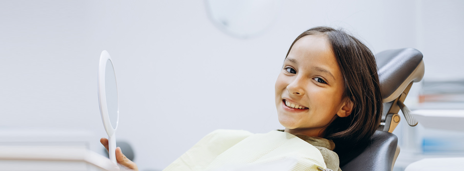 The image is a photograph of a young child sitting in a dental chair, smiling at the camera.