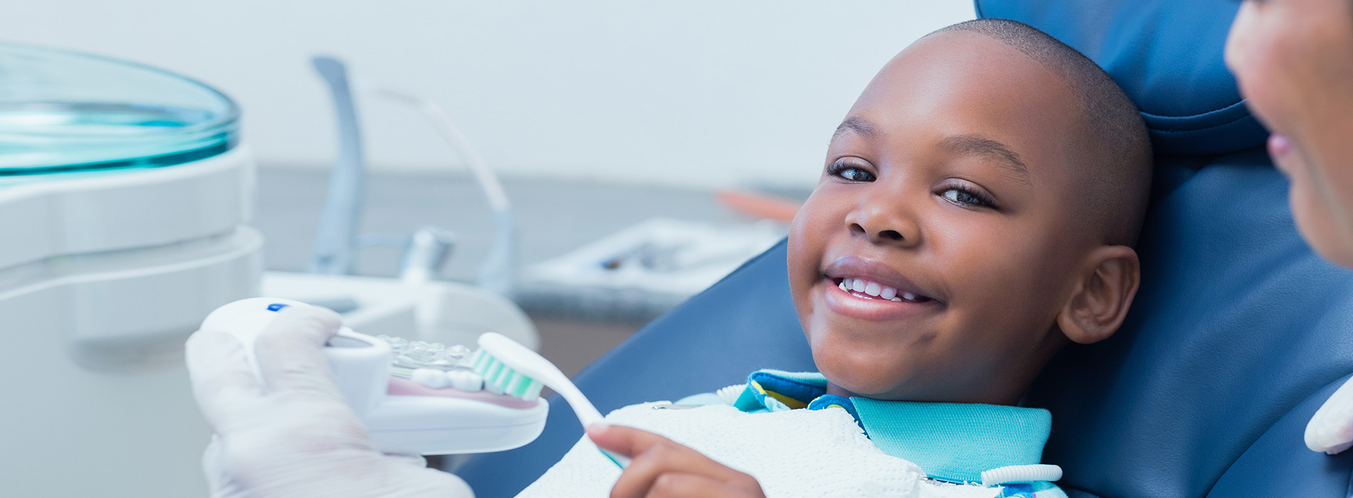 A young boy in a dental chair, smiling and looking at the camera, with a dentist sitting behind him.