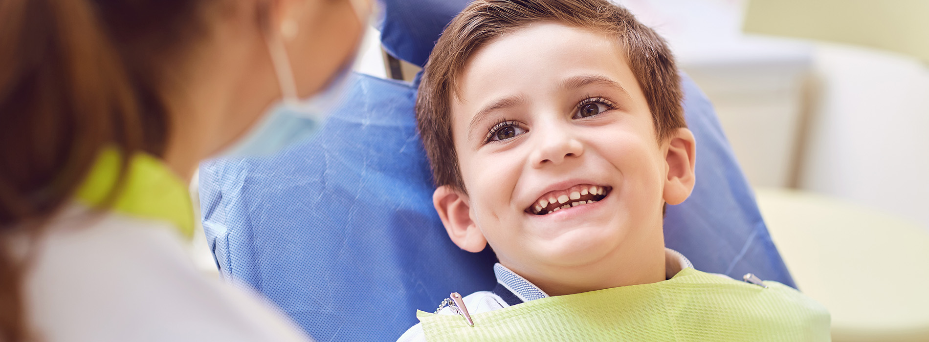 The image shows a young child sitting in a dental chair, smiling and looking towards the camera, with a dentist standing behind them, wearing a white coat and engaging in conversation.