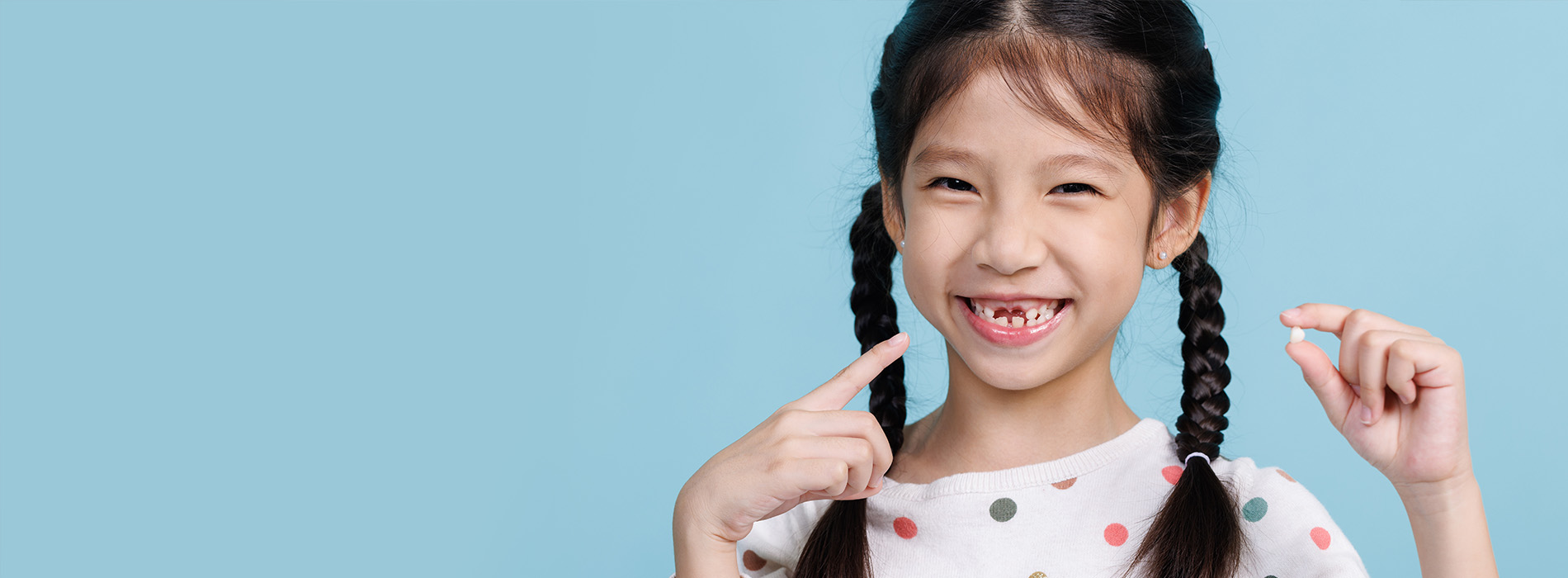 The image shows a young girl with a joyful expression, making a playful gesture with her hands near her face. She is standing against a blue background.