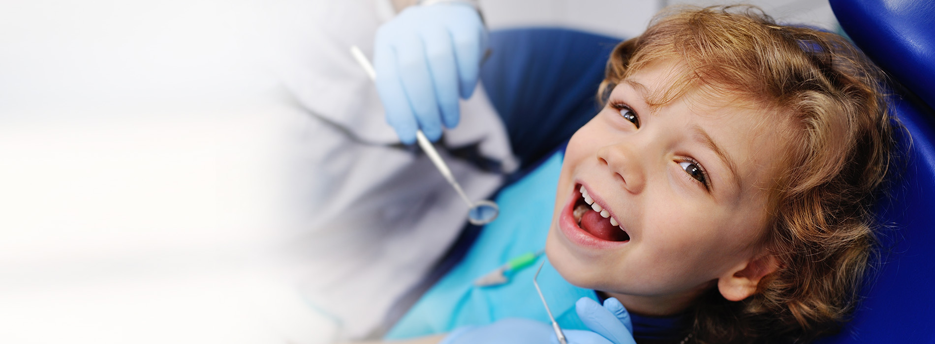 A young child is smiling and holding a toothbrush, sitting in a dental chair with a backdrop of medical equipment.