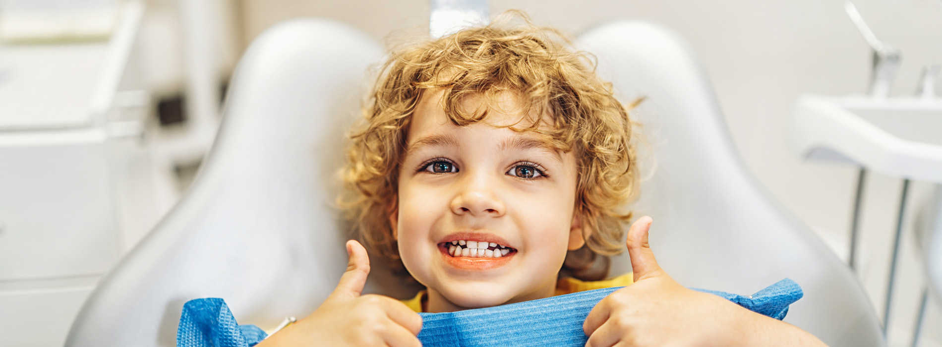 Young child in dental chair, giving thumbs up.