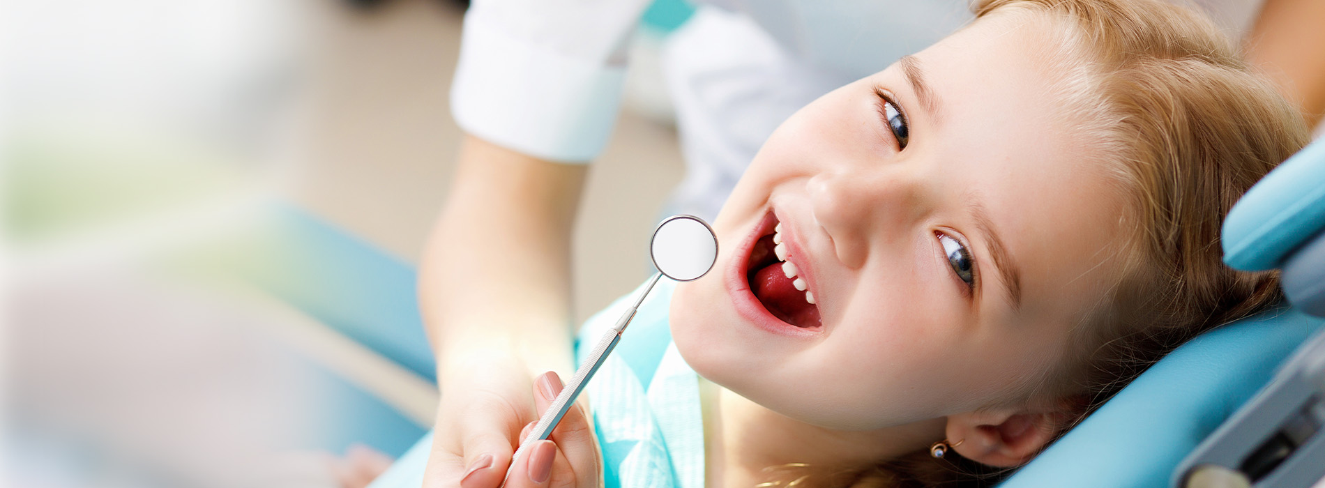 A young child is seated in a dental chair, smiling and looking directly at the camera, with a dental professional holding a mirror to their side.